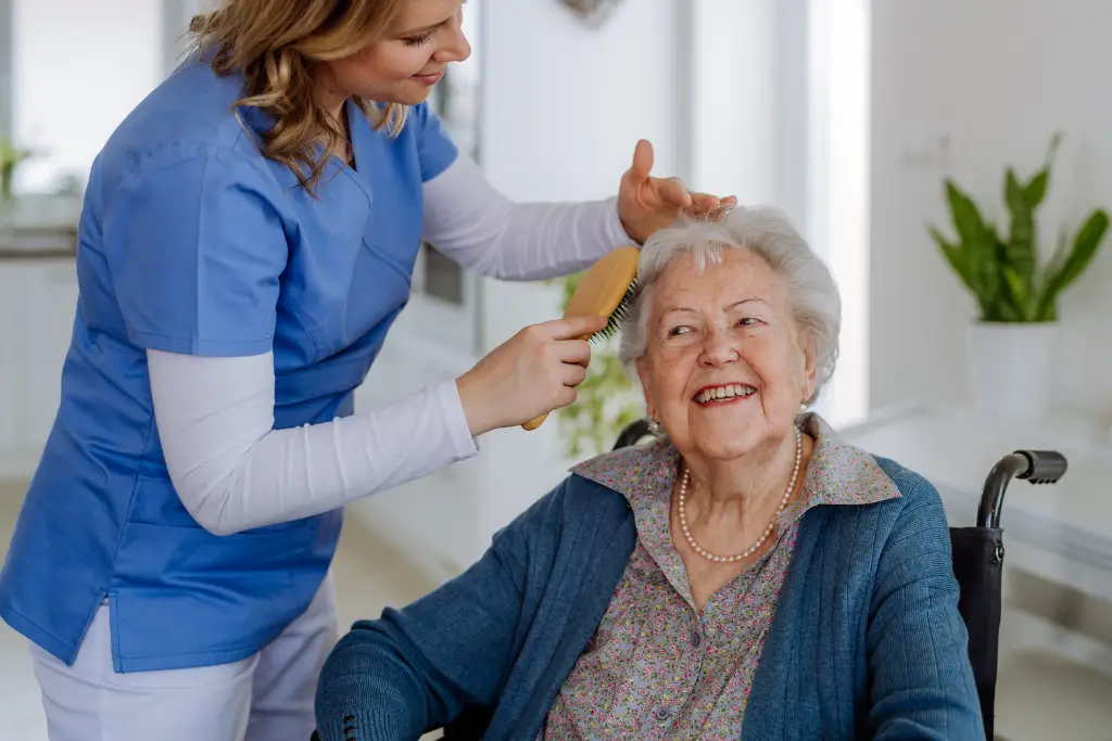 Cuidadora del personal sonríe mientras cepilla el pelo de una residente adulta mayor feliz en silla de ruedas